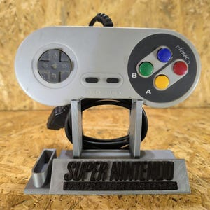 Puede incluir: Un mando de videojuegos gris claro con un pad direccional circular, dos botones ovalados pequeños etiquetados como Select y Start, y cuatro botones de acción de colores etiquetados como A, B y Turbo. El mando descansa sobre un soporte gris con la etiqueta Super Nintendo.