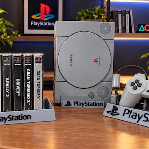 Pode incluir: Uma consola de jogos Sony PlayStation cinzenta, um comando cinzento a condizer e seis caixas de jogos, incluindo Toca 2 Touring Cars, Colin McRae Rally, V-Rally 2, Driver e dois títulos Gran Turismo 2, expostos em suportes cinzentos com o logótipo PlayStation.