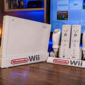 Pode incluir: Uma consola de jogos Nintendo Wii branca numa base, acompanhada por uma estação de carregamento branca com dois comandos Wii Remote e dois controladores Nunchuk. Tanto a base da consola como a estação de carregamento exibem o logótipo Nintendo Wii.