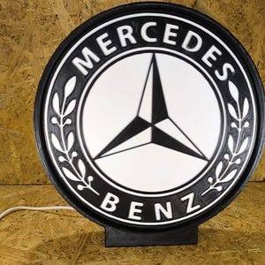 Könnte beinhalten: Rundes Mercedes-Benz-Schild in Schwarz und Weiß mit dem Markenlogo in der Mitte. Das Schild hat einen schwarzen Rand mit den weißen Worten "MERCEDES" und "BENZ". Es ist auf einem schwarzen Sockel montiert.