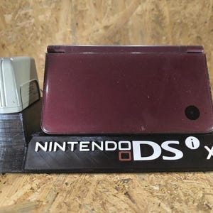 Può includere: Una console di gioco portatile Nintendo DSi XL color bordeaux appoggiata su un supporto espositivo nero stampato in 3D. Il supporto presenta la scritta bianca NINTENDO DSi XL sul davanti. Un alimentatore grigio chiaro è posizionato a sinistra.