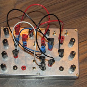 Könnte beinhalten: Eine silberne Metallbox mit mehreren Knöpfen, Schaltern und Buchsen. Rote, schwarze und weiße Drähte sind an den Buchsen angeschlossen. Die Box steht auf einer Holzoberfläche.