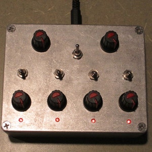 Puede incluir: Una caja de metal plateada con cuatro perillas negras, cuatro LED rojos y cuatro interruptores de palanca. La caja tiene un interruptor de encendido negro y un cable negro.