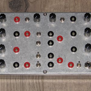 May include: A silver metal panel with black knobs, red and black buttons, and silver switches. The panel is a music synthesizer module.