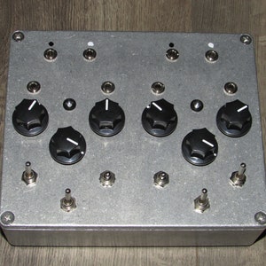 Puede incluir: Una caja de metal plateada con seis botones negros, cuatro interruptores de palanca y seis tornillos plateados. La caja tiene una superficie texturizada.