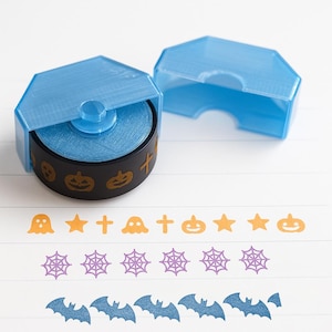 Könnte beinhalten: Ein blauer Klebebandabroller mit einer schwarzen Spule mit goldenen Halloween-Designs. Der Abroller ist geöffnet und zeigt das Klebeband. Darunter befinden sich dekorative Bänder mit Halloween-Motiven: Geister, Sterne, Kreuze, Kürbisse, Spinnennetze und Fledermäuse.