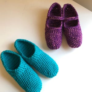 May include: Two pairs of handmade crochet slippers. One pair is a vibrant teal, the other is a deep purple with a strap across the top. The slippers are made with a thick yarn, suggesting warmth and comfort.