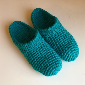 May include: A pair of teal crochet slippers. The slippers are made of a thick yarn, with a textured appearance. The slippers are designed to cover the foot and ankle.