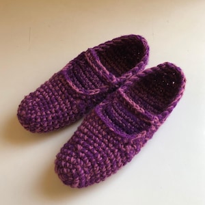 May include: A pair of handmade, crocheted slippers in shades of purple and pink. The slippers have a closed-toe design with a strap across the top of the foot. The yarn appears to be a soft, textured material, perfect for indoor wear.
