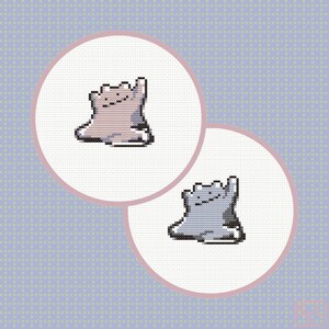 May include: Two circular cross-stitch patterns on white fabric, each featuring a pixelated, blob-like creature with a simple face. One creature is rendered in shades of light pink and purple, while the other is in shades of light blue and gray.