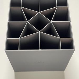 以下が含まれることがあります： 幾何学模様のグレーのプラスチック製オーガナイザーで、16個のコンパートメントがあります。オーガナイザーには、底面に「VITRA」という文字が印刷されています。