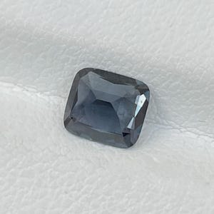 Puede incluir: Una gema tallada cuadrada, facetada, de color azul ahumado intenso. La gema tiene forma de coj&iacute;n con esquinas redondeadas y una superficie pulida, que refleja la luz. La piedra est&aacute; sobre un fondo blanco.