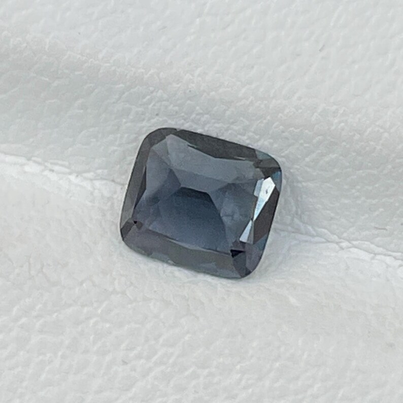 Puede incluir: Una gema tallada cuadrada, facetada, de color azul ahumado intenso. La gema tiene forma de coj&iacute;n con esquinas redondeadas y una superficie pulida, que refleja la luz. La piedra est&aacute; sobre un fondo blanco.