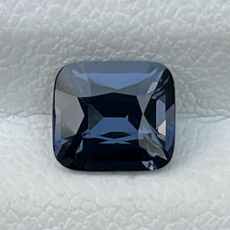 Puede incluir: Una gema tallada cuadrada y facetada, de color azul intenso. La superficie pulida de la gema refleja la luz, creando sombras y reflejos. La gema est&aacute; sobre un fondo blanco texturizado.