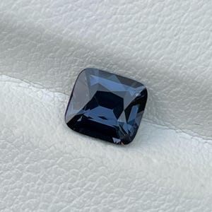 Puede incluir: Una gema azul oscuro de corte cuadrado con una superficie pulida. La gema refleja la luz, mostrando facetas y profundidad interna. La piedra se encuentra sobre un fondo blanco texturizado, resaltando su color y claridad. Esta gema es adecuada para joyer&iacute;a.