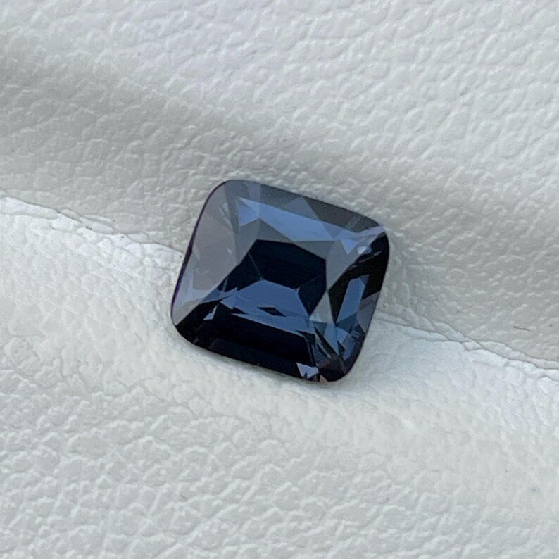 Puede incluir: Una gema azul oscuro de corte cuadrado con una superficie pulida. La gema refleja la luz, mostrando facetas y profundidad interna. La piedra se encuentra sobre un fondo blanco texturizado, resaltando su color y claridad. Esta gema es adecuada para joyer&iacute;a.