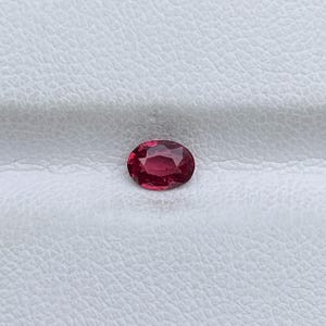 Può includere: Una gemma rossa sfaccettata di forma ovale. La gemma è di un colore rosso intenso e ricco e sembra ben lucidata, riflettendo la luce. La gemma è posta su uno sfondo bianco strutturato.