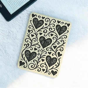 Puede incluir: Una funda de lector electrónico de color crema con un diseño de corazón y vid negro. La funda tiene forma rectangular y está decorada con grandes corazones y pequeños acentos de corazón. Un lector electrónico es visible en el fondo.