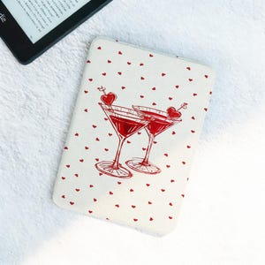 May include: A white Kindle cover with a pattern of small red hearts and two red martini glasses. Each glass has a heart-shaped garnish. The cover is rectangular and has rounded corners.