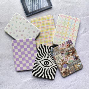 Puede incluir: Seis fundas rectangulares para lector electrónico con varios diseños, incluyendo estrellas pastel, cuadros multicolores, tablero de ajedrez morado, un patrón de ojo con rayos negros y blancos, y una ilustración de librería con el texto BOOK STORE.