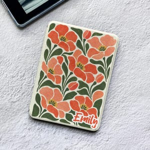 May include: A rectangular e-reader case featuring a cream background with a repeating pattern of orange flowers and dark green leaves. The name Emily is printed in a stylized orange font in the bottom right corner of the protective cover.
