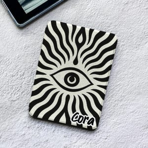 Puede incluir: Una funda rectangular para lector electrónico con un estampado gráfico en blanco y negro de un ojo central rodeado de líneas onduladas radiantes. El nombre Cora está impreso en una fuente cursiva negra en la esquina inferior derecha.