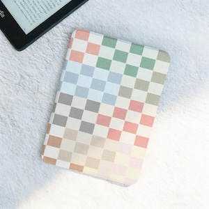 May include: A pastel-colored checkerboard pattern Kindle cover. The cover features squares in shades of pink, green, blue, gray, and beige. A black Kindle device is partially visible in the upper left corner, displaying text on the screen.