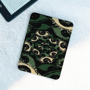 May include: A rectangular e-reader cover featuring a repeating eye pattern in shades of green, black, and cream. The cover is placed on a white surface, with a portion of an e-reader visible in the upper left corner.