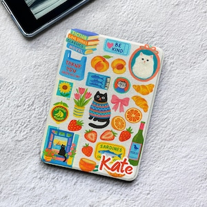 Puede incluir: Una funda rectangular para lector electrónico con fondo blanco cubierta de pegatinas coloridas que incluyen una pila de libros, una bolsa azul, un gato negro con suéter, fresas, naranjas, cruasanes, una botella de vino y el nombre Kate.
