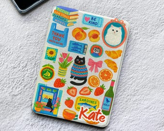 May include: A rectangular e-reader case featuring a white background covered in colorful stickers including a stack of books, a blue thank you bag, a black cat in a sweater, strawberries, oranges, croissants, a wine bottle, and the name Kate.
