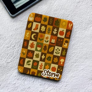 May include: A rectangular e-reader cover featuring a chequered pattern of autumn-themed icons including leaves, pumpkins, apples, mushrooms, flowers, a candle, a crescent moon, and a coffee cup. The name Storm is printed in white script at the bottom.