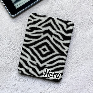 May include: A rectangular electronic reader cover featuring a black and white zebra stripe pattern with a diamond-shaped center design. The word Hero is printed in a white, stylized, bold font with a black outline in the bottom right corner.