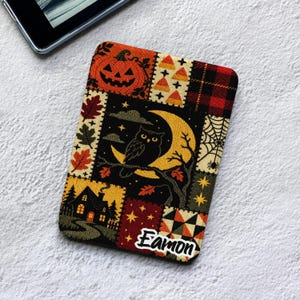 May include: A rectangular e-reader cover featuring a patchwork design with Halloween motifs including a jack-o'-lantern, candy corn, a spider web, a haunted house, and an owl perched on a crescent moon. The name Eamon is printed in white.