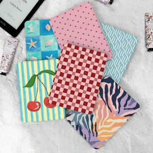 May include: A selection of patterned book covers in various designs. Includes pink stars, red checkered, blue and white wave, and a cherry illustration. A Kindle e-reader and hand cream are also visible.