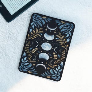 Könnte beinhalten: Schwarze, rechteckige E-Reader-Hülle mit einem Himmelsdesign. Die Hülle zeigt Mondphasen in Weiß und Hellblau, umgeben von goldenen und blauen botanischen Illustrationen. Ein Kindle-E-Reader ist teilweise oben links zu sehen.