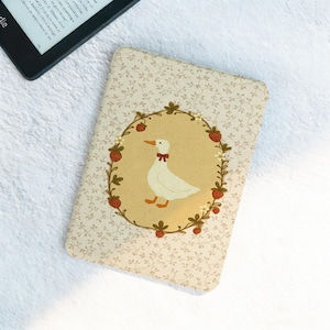 Puede incluir: Una funda Kindle de color crema con un estampado floral y una ilustración central de un ganso blanco con una pajarita roja, rodeado por una corona de fresas y hojas. La funda es rectangular y está diseñada para proteger un lector electrónico.