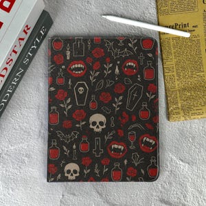 May include: A black tablet case with a repeating pattern of red roses, vampire fangs, skulls, potion bottles, and coffins. A white stylus rests on the case. The case is next to two books and a newspaper.