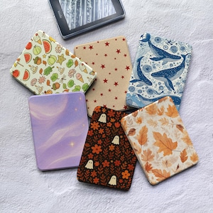 May include: Six rectangular e-reader cases featuring various designs: one with colorful autumn icons, one with red stars on tan, one with blue whales, one with a purple nebula, one with white ghosts on a dark floral background, and one with orange leaves.