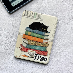 Puede incluir: Una funda rectangular para lector electrónico con un gato negro descansando sobre una pila de cinco libros coloridos. Debajo de los libros hay una taza de té sobre un platillo con una mancha. El nombre Fran está impreso en letras negras abajo.