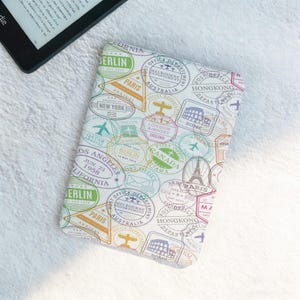 Puede incluir: Una funda blanca para Kindle con un diseño de sellos de viaje, que incluye sellos de Berlín, París y Nueva York. La funda presenta sellos coloridos e ilustraciones de aviones. Un lector Kindle es parcialmente visible en el fondo.