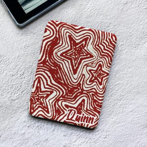 Może przedstawiać: Prostokątne etui na czytnik e-booków z powtarzającym się wzorem czerwonych i białych falistych gwiazd. Projekt zawiera imię Quinn nadrukowane czerwoną czcionką skryptową w prawym dolnym rogu etui, które ma gładką powierzchnię.