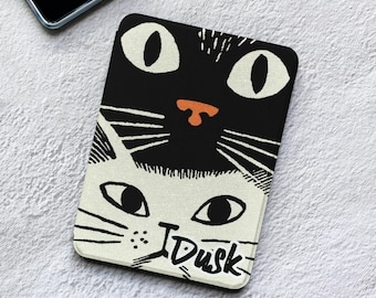 Puede incluir: Una funda rectangular para lector electrónico con una ilustración gráfica de dos caras de gato. El gato superior es negro con grandes ojos blancos y nariz naranja, el inferior es blanco con ojos negros. La palabra Dusk está impresa en negro.