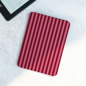May include: A rectangular Kindle cover with a vertical striped pattern in shades of red and pink. The cover is placed on a white, textured surface next to a black e-reader.