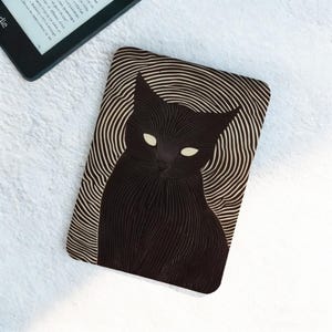 Puede incluir: Funda Kindle con diseño de gato negro. La funda presenta una ilustración de un gato negro con ojos blancos brillantes, sobre un fondo de líneas onduladas concéntricas. La funda rectangular probablemente está hecha de un material duradero.