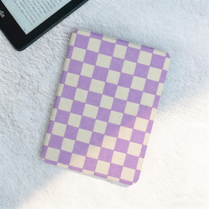 May include: A rectangular Kindle cover with a checkerboard pattern in lavender and off-white. The cover is closed and lies on a white, textured surface. A black Kindle device is partially visible in the upper left corner.