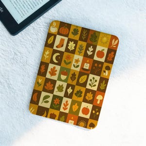 May include: A rectangular, fall-themed case with a checkerboard pattern of autumn icons, including leaves, pumpkins, and candles. The case is in shades of brown, orange, and green. A Kindle e-reader is visible in the upper left corner.