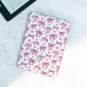 May include: White Kindle cover featuring a pattern of pink bows and hearts. The rectangular cover is made of fabric. A black Kindle device is partially visible in the upper left corner.