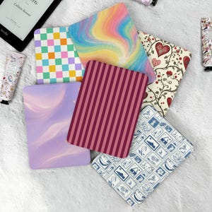 Puede incluir: Una colección de coloridas fundas para Kindle con varios diseños, incluyendo un patrón de tablero de ajedrez, un arcoíris arremolinado y motivos florales. También se ven un lector Kindle y tubos de crema de manos.