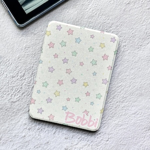 May include: A rectangular e-reader case featuring a white background with a repeating pattern of small pastel-colored stars in pink, blue, yellow, and green. The name Bobbi is printed in a light pink script font in the bottom right corner.