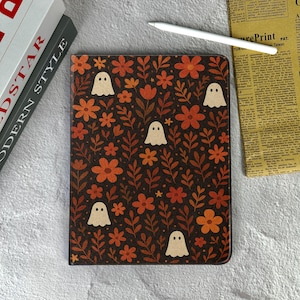 May include: A dark brown notebook cover with a floral and ghost pattern. The design features orange flowers, brown leaves, and small white ghosts. A white stylus and a stack of books are visible in the background.